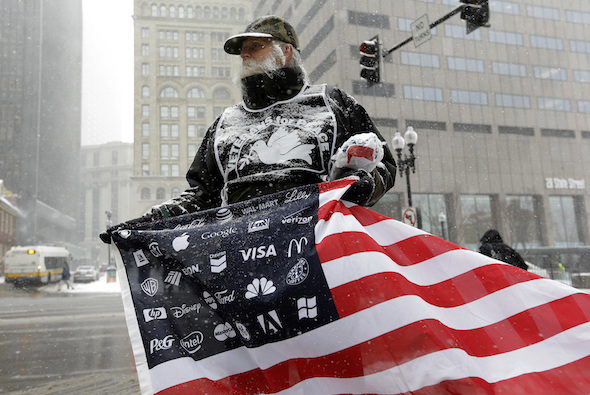 Protester Bob Bowes, of Somerville, Mass., displays an American flag featuring corporate logos outside a meeting of U.S. elites in Boston. (Steven Senne / AP)