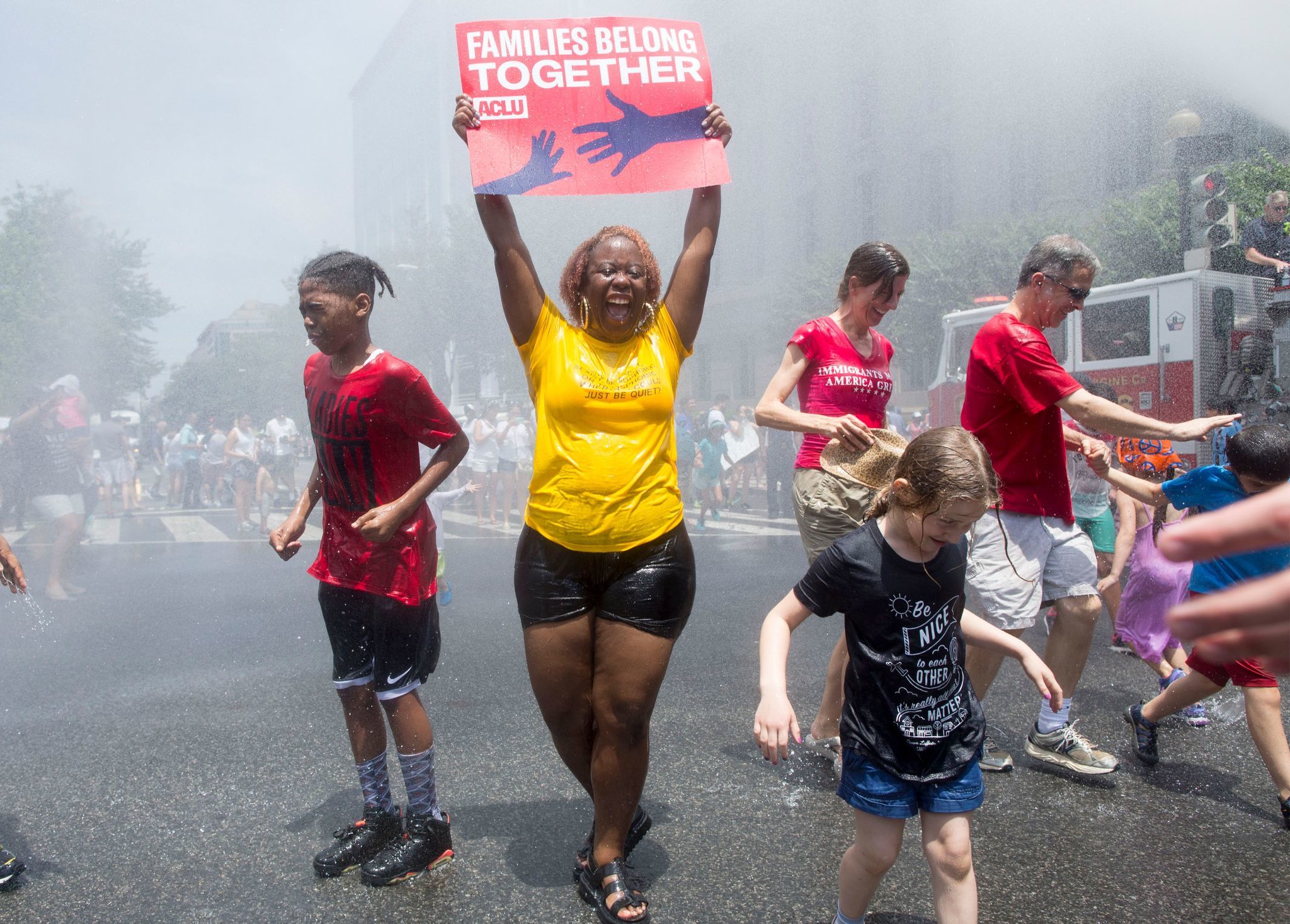 WASHINGTON, D.C.: A woman holds a sign while joining others underneath water being sprayed by a firetruck to cool off people attending a Families Belong Together rally. Michael Reynolds / EPA / Shutterstock